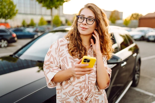 Woman in glasses smiling at her phone beside a car